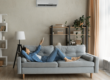 Woman relaxing on couch pointing remote at AC wall unit